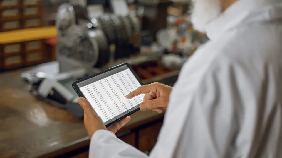 Illustrative photo: Worker reviewing a table of numbers on a tablet beside industrial equipment.
