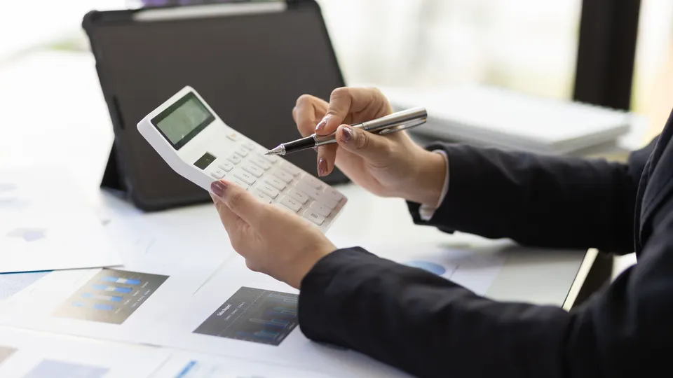 Illustrative photo: Person checking figures with a calculator and printed charts.