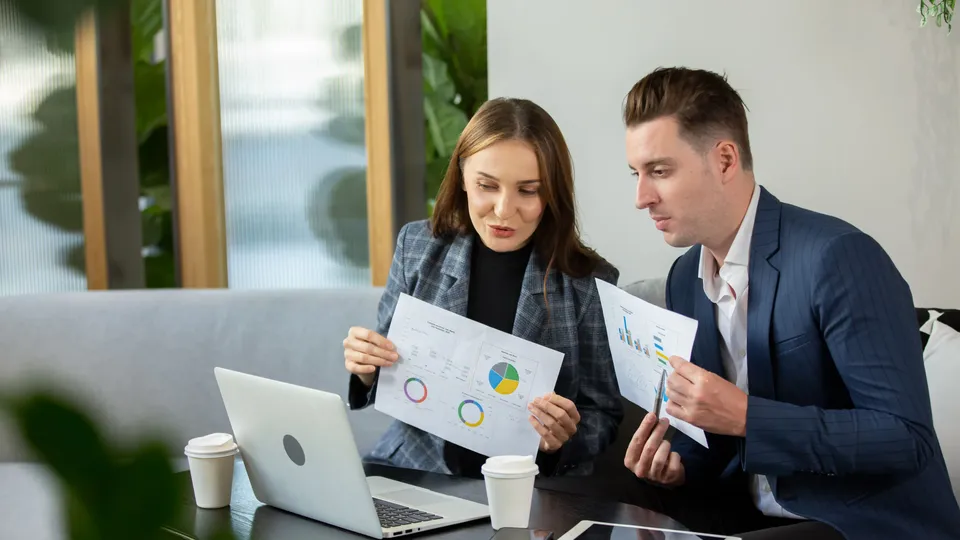 Illustrative photo: Two colleagues comparing printed charts beside a laptop.
