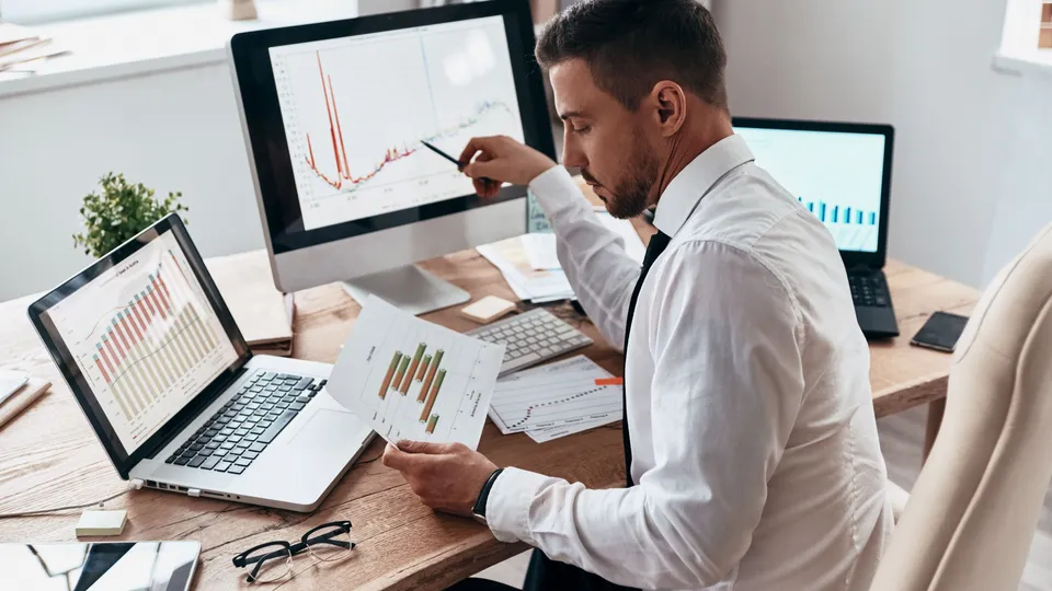 Illustrative photo: Analyst reviewing charts on a laptop, monitor and printed report.