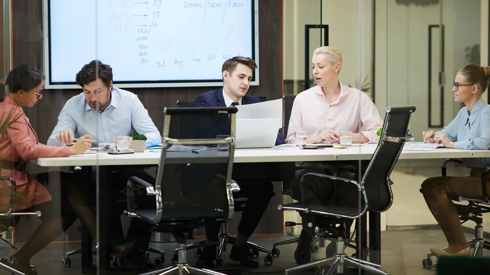 Illustrative photo: Team meeting around a table with laptops and a presentation screen.