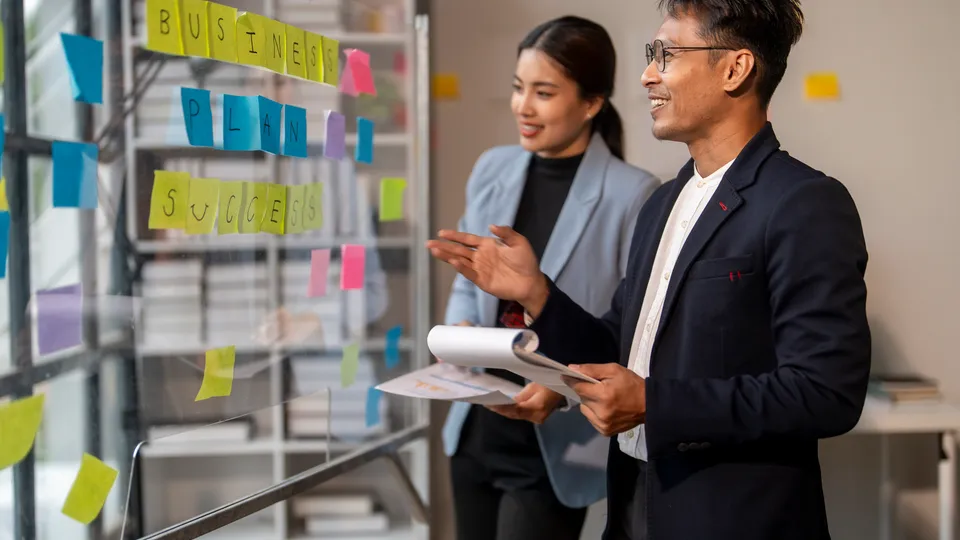 Illustrative photo: Two colleagues discussing sticky notes on a glass wall.