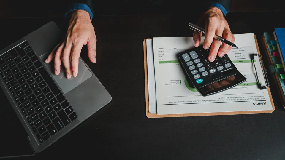 Illustrative photo: Hands using a calculator beside a laptop and financial paperwork.