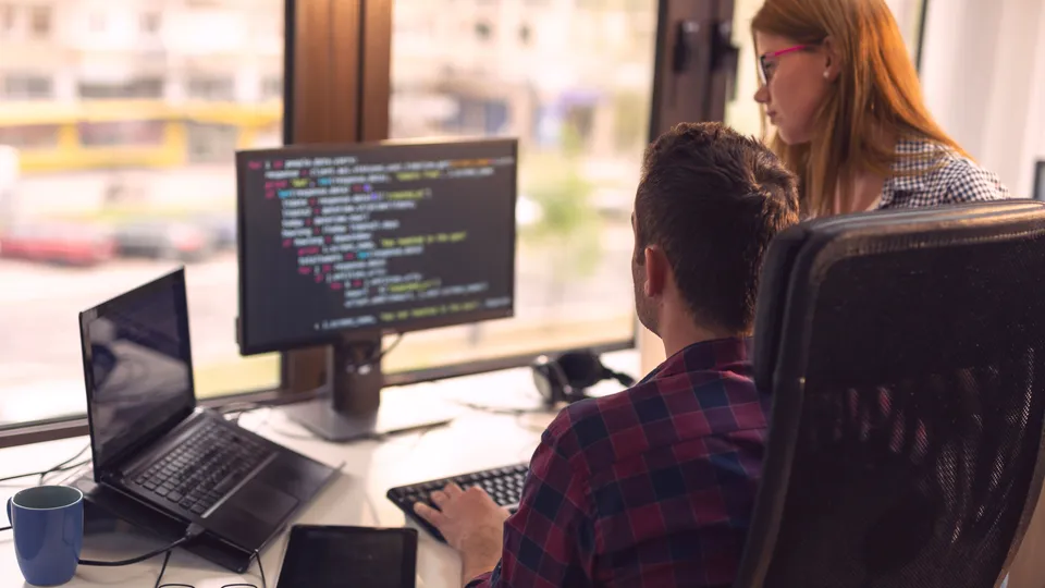 Illustrative photo: Two developers reviewing source code at a workstation.