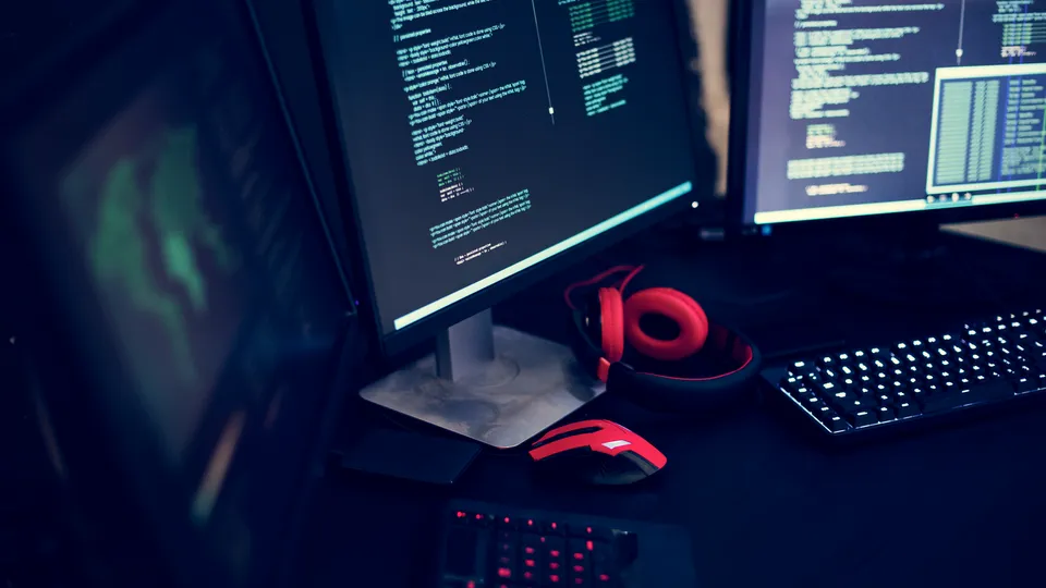Illustrative photo: Source code on multiple monitors beside headphones and a keyboard.