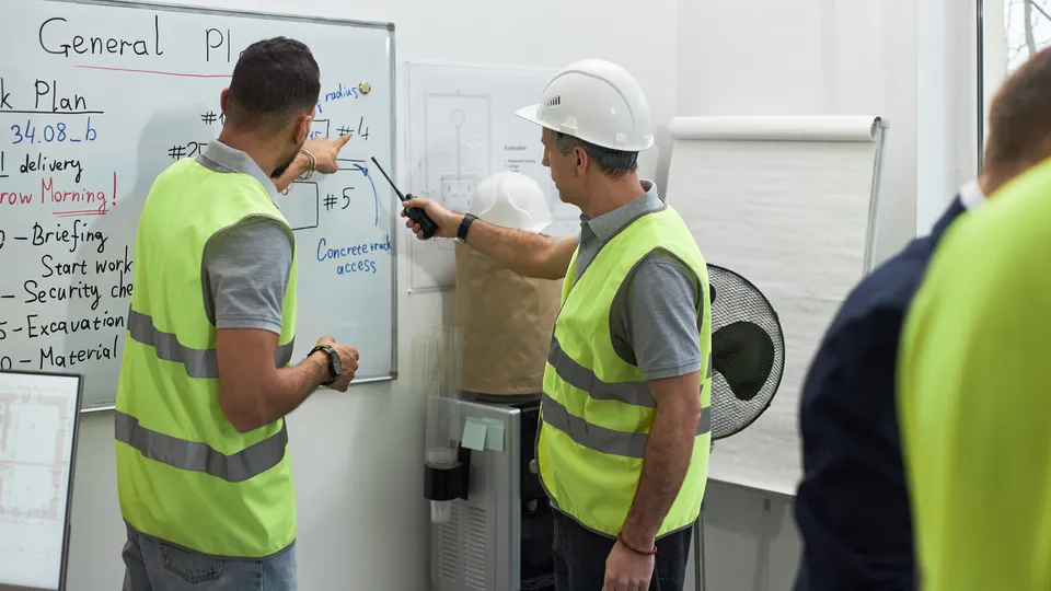 Illustrative photo: Workers in high-visibility vests discussing a plan on a whiteboard.