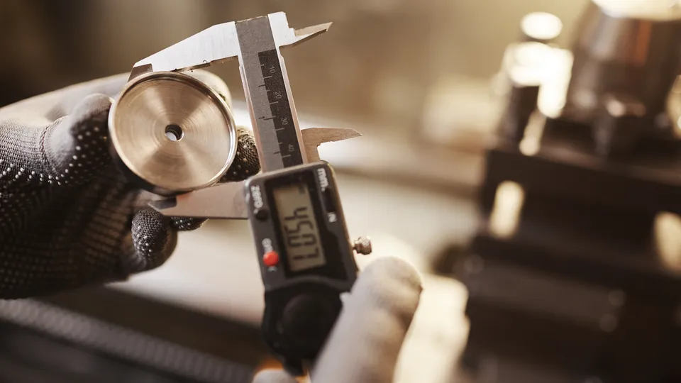 Illustrative photo: Worker measuring a metal component with a digital caliper.