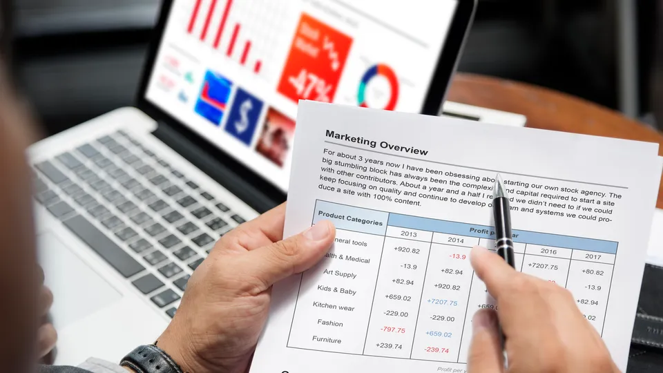 Illustrative photo: Hands comparing a marketing report with charts on a laptop.