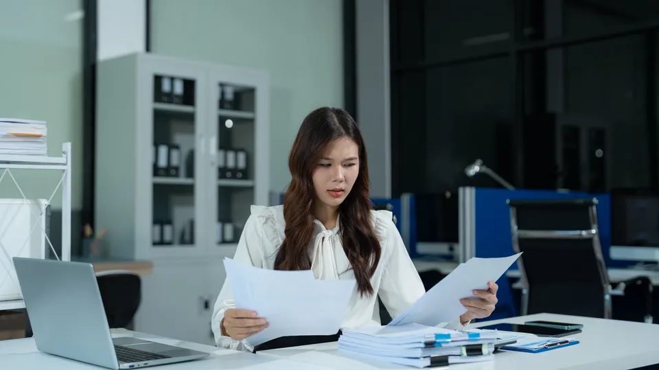 Illustrative photo: Woman checking paperwork beside a calculator and laptop.