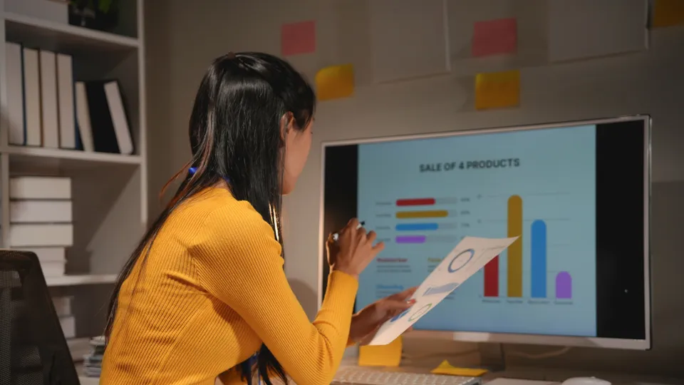 Illustrative photo: Woman comparing printed charts with a sales report on a monitor.