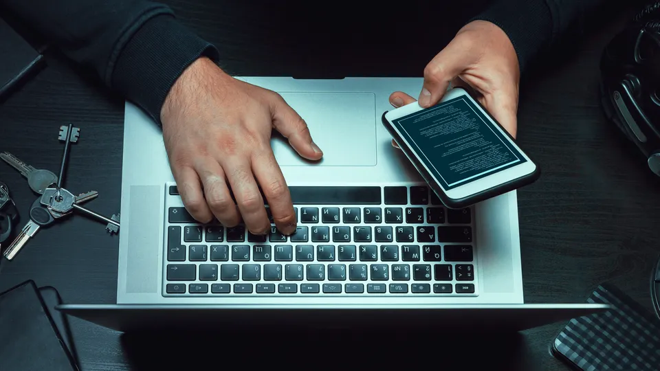 Illustrative photo: Person working with a laptop and a phone displaying code.