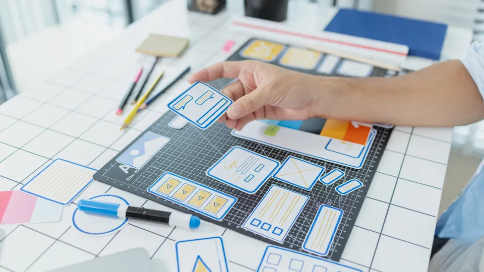 Illustrative photo: Hand holding a paper wireframe over interface design sketches.