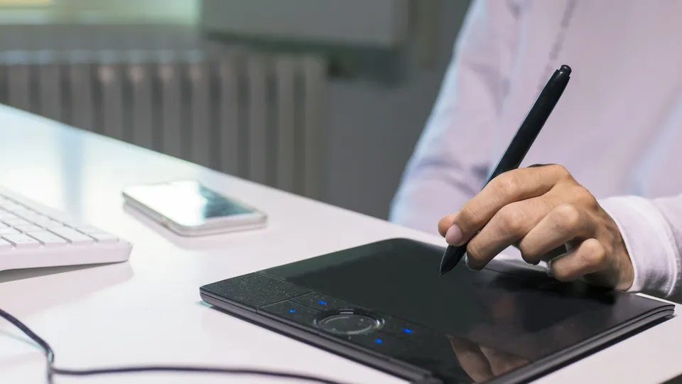 Illustrative photo: Hand using a stylus on a graphics tablet beside a keyboard.