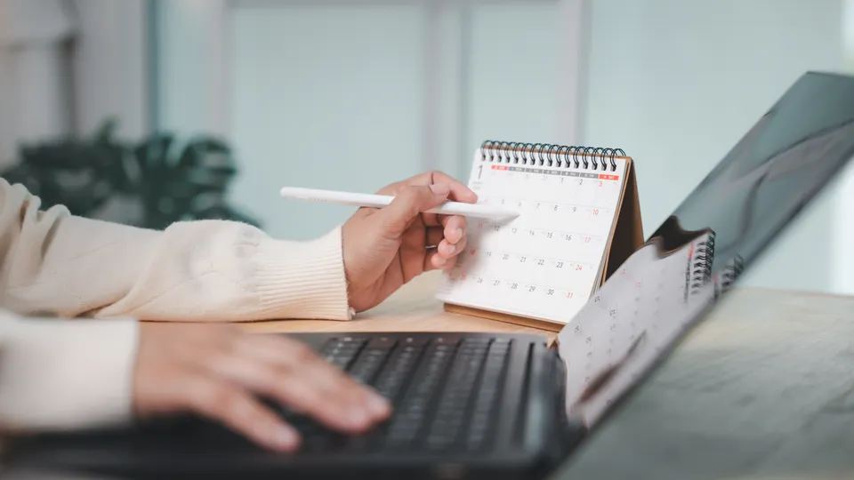 Illustrative photo: Hands writing in a desk calendar beside a laptop.
