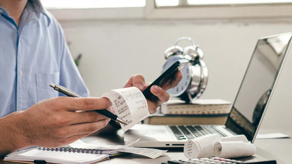 Illustrative photo: Person checking receipts with a calculator beside a laptop.