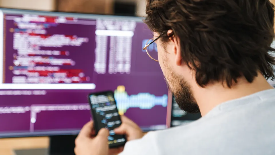 Illustrative photo: Developer using a smartphone beside a monitor displaying code.