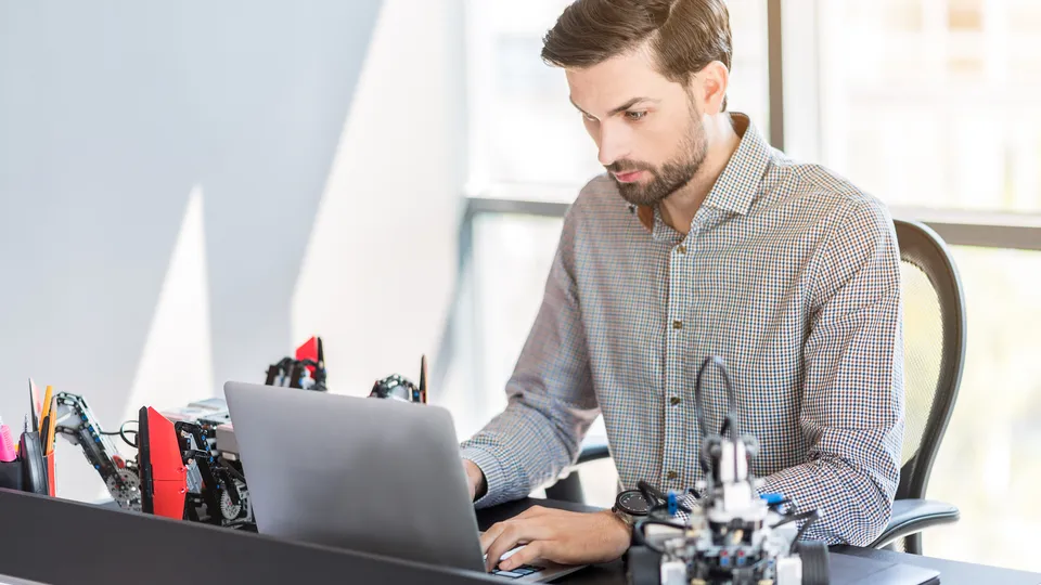 Illustrative photo: Man working on a laptop beside small robot models.