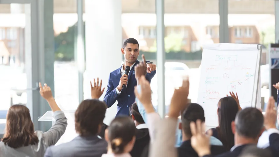 Illustrative photo: Presenter addressing an audience with raised hands.