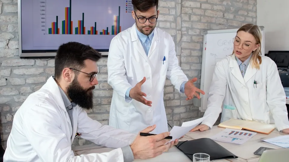Illustrative photo: Researchers in lab coats discussing reports beside a chart display.