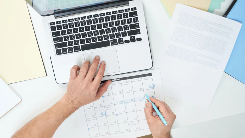 Illustrative photo: Person planning dates on a paper calendar beside a laptop.
