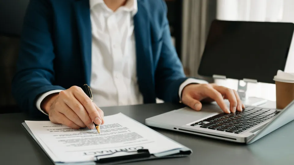 Illustrative photo: Person signing a document beside a laptop.