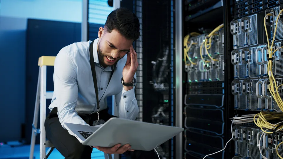 Illustrative photo: Administrator holding a laptop beside server racks.