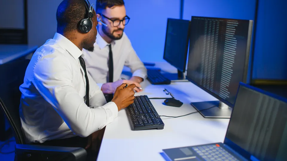Illustrative photo: Two colleagues reviewing code on desktop monitors.