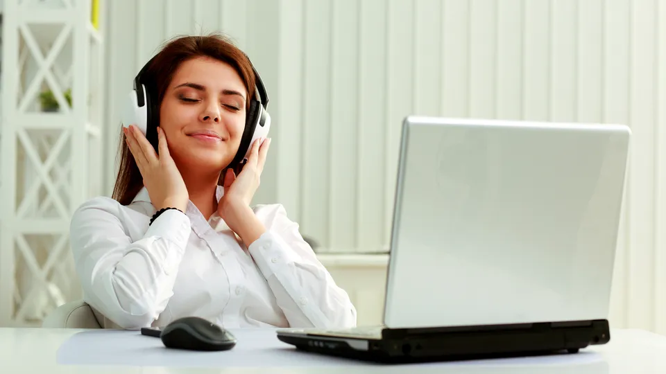 Illustrative photo: Woman listening to music with headphones at a desk.