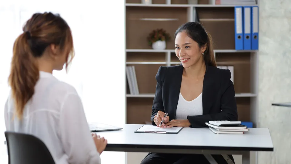 Illustrative photo: Interviewer speaking with an applicant across an office desk.