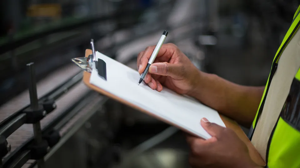 Illustrative photo: Worker recording notes on a clipboard in a factory.
