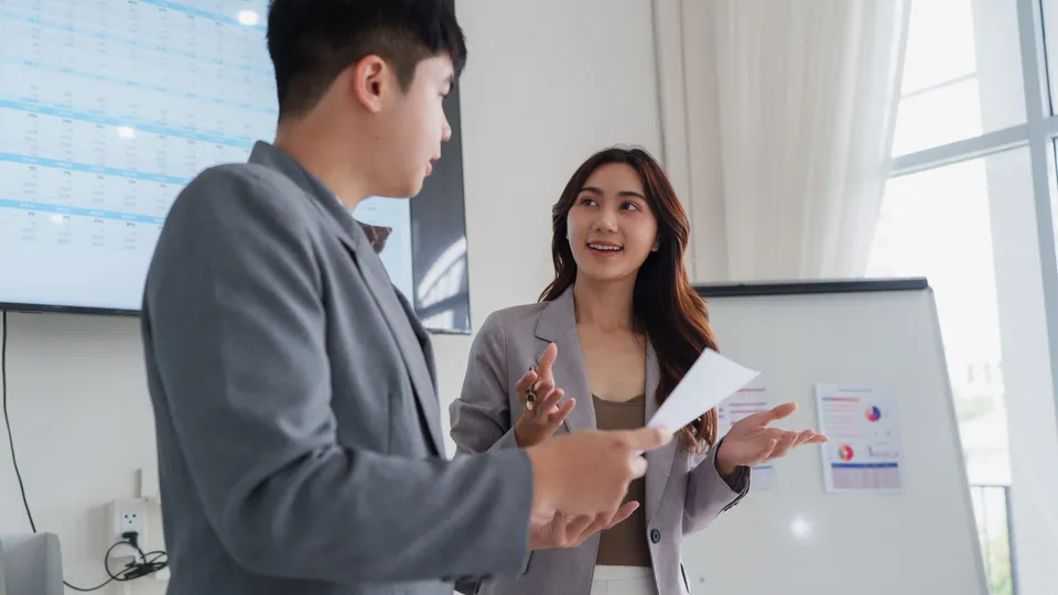 Illustrative photo: Two professionals discussing charts beside a display.